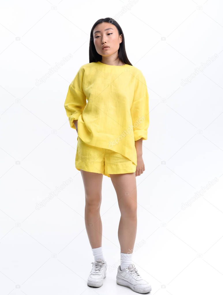 Beautiful Asian girl in a yellow linen casual suit posing against a white wall in a photo studio