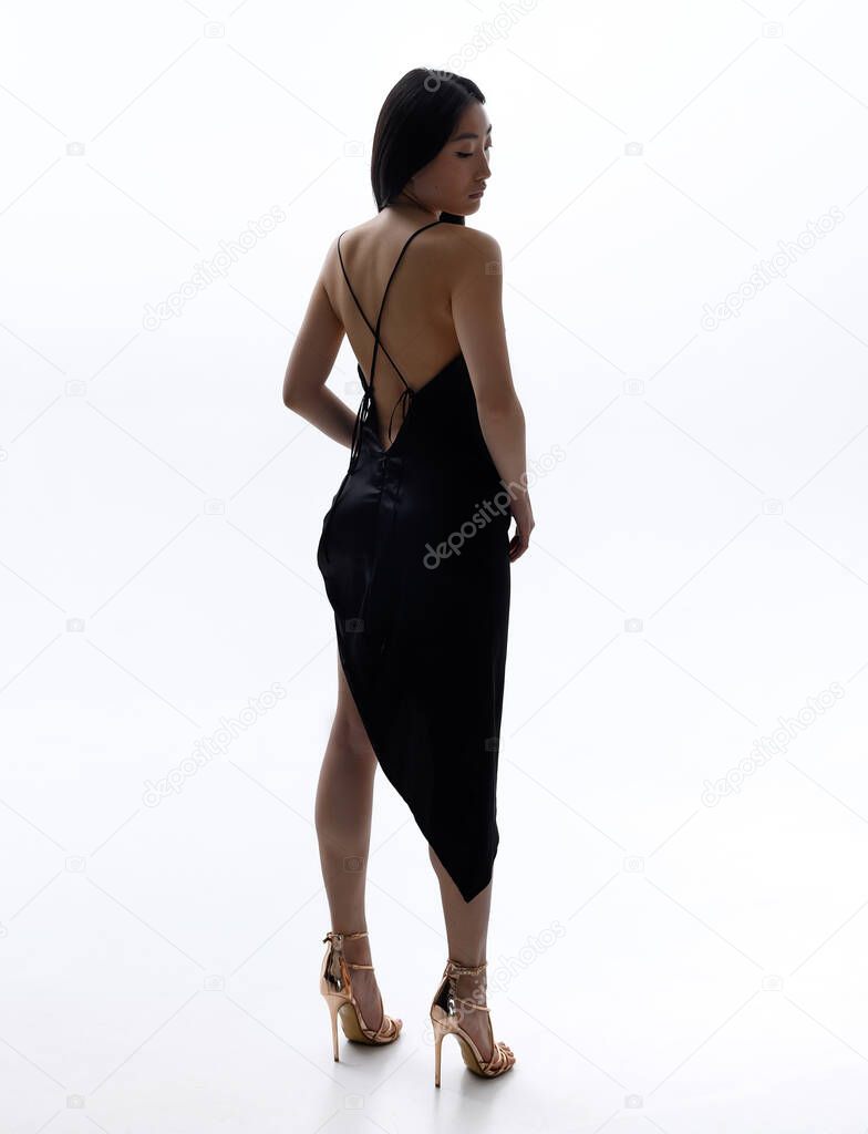 A beautiful Asian girl in a black dress is posing against a white wall in a photo studio. Fashion shooting