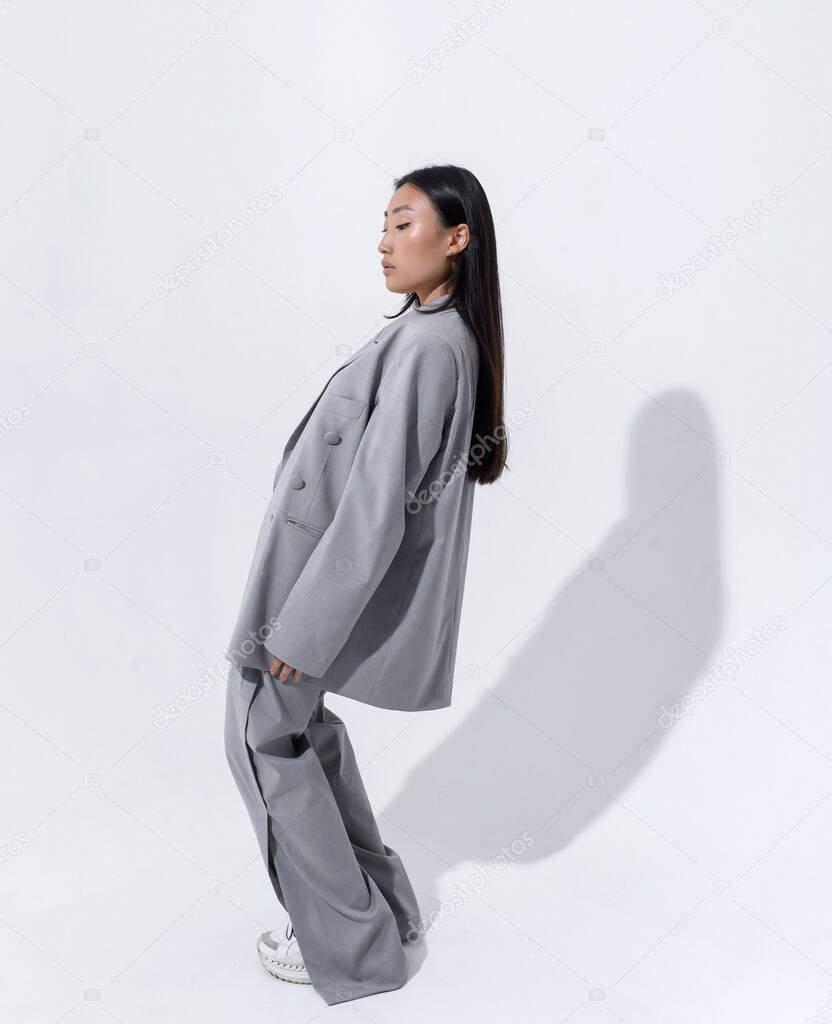 A beautiful Asian girl in a gray suit on a naked body poses against a white wall in a photo studio. Fashion shooting