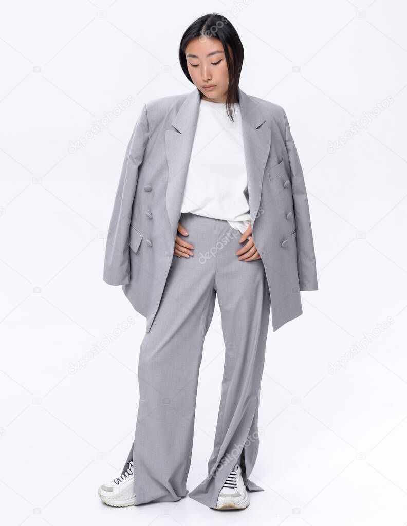 A beautiful Asian girl in a gray suit poses against a white wall in a photo studio. Fashion shooting