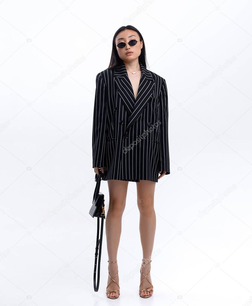 Beautiful Asian girl in black striped jacket wearing sunglasses posing against white wall in photo studio. Fashion shooting