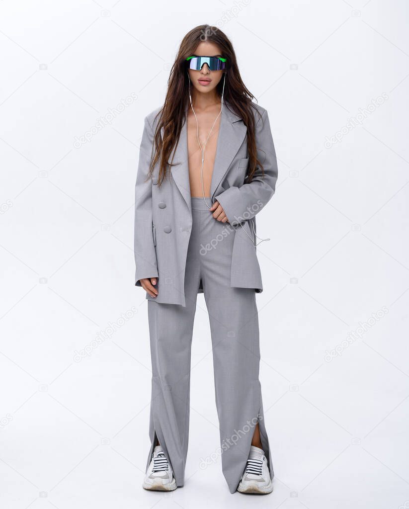 Beautiful Caucasian brunette girl in stylish glasses in a gray fashion suit posing on a white background in headphones