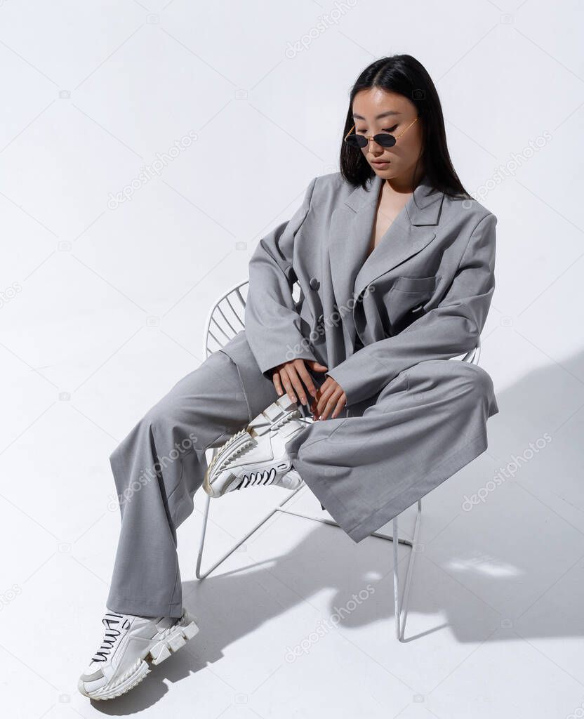 A beautiful Asian girl in a gray suit on a naked body poses against a white wall in a photo studio. Fashion shooting