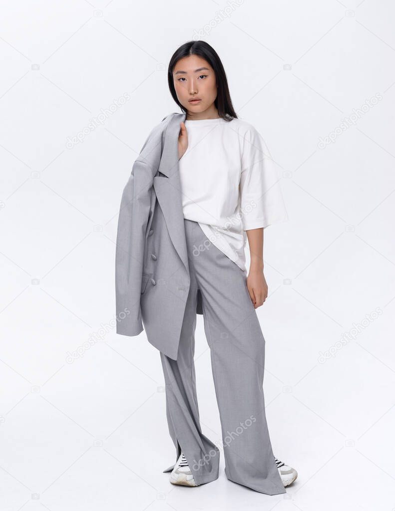 A beautiful Asian girl in a gray suit poses against a white wall in a photo studio. Fashion shooting
