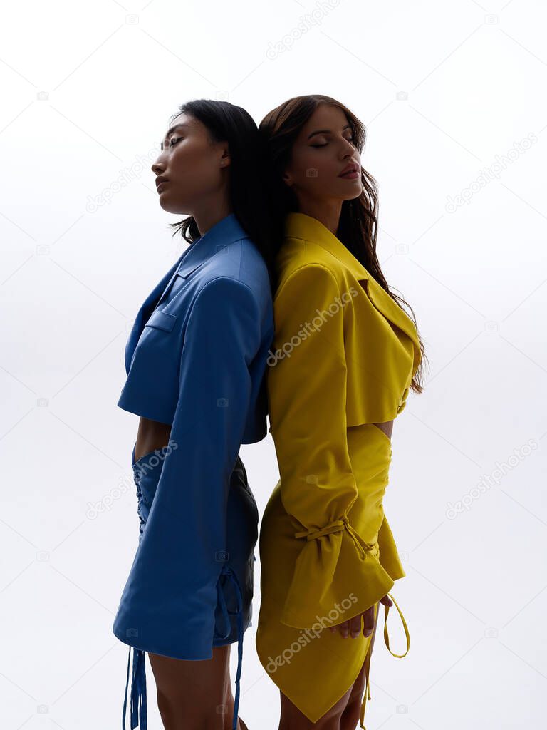 Two beautiful Caucasian and Asian girls lie on the white floor and pose in blue and yellow suits in a photo studio