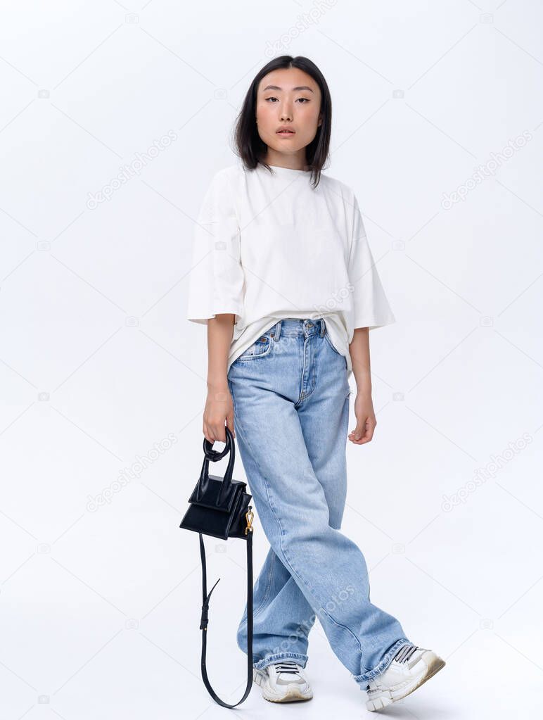 A beautiful Asian girl in a white t-shirt and blue jeans poses against a white wall in a photo studio. Fashion shooting