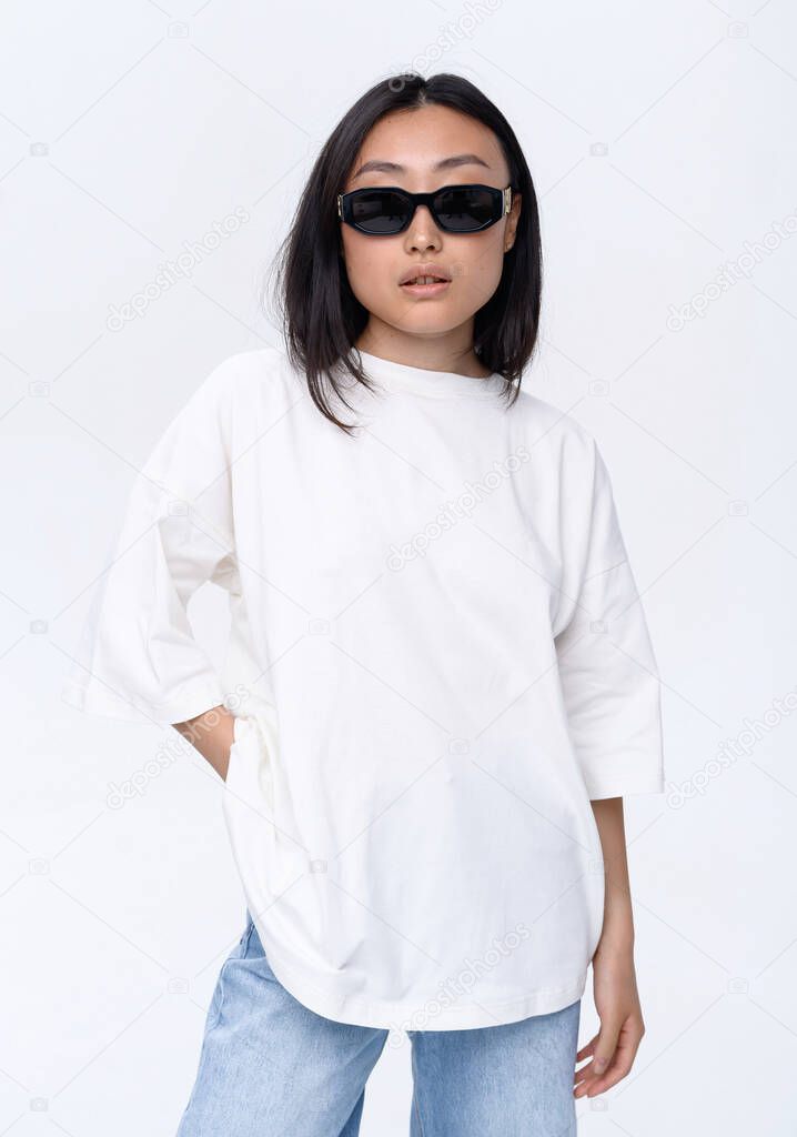 A beautiful Asian girl in a white t-shirt and blue jeans poses against a white wall in a photo studio. Fashion shooting