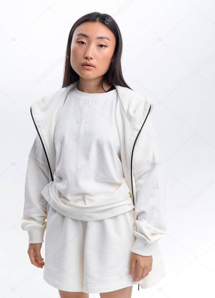 A beautiful Asian girl in a white casual tracksuit poses against a white wall in a photo studio