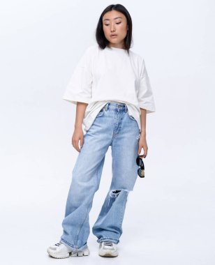 Beyaz tişörtlü ve kot pantolonlu güzel bir Asyalı kız fotoğraf stüdyosunda beyaz bir duvara karşı poz veriyor. Moda çekimi.