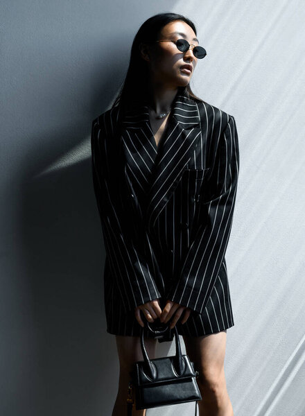 Beautiful Asian girl in black striped jacket wearing sunglasses posing against gray wall in photo studio. Fashion shooting