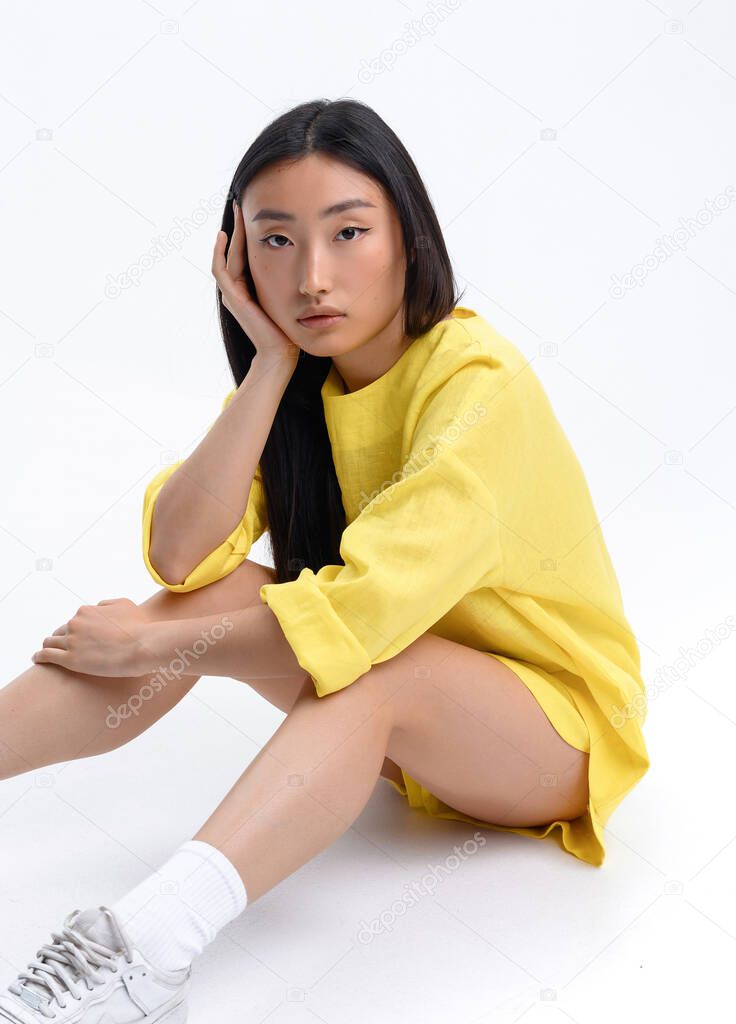 Beautiful Asian girl in a yellow linen casual suit posing against a white wall in a photo studio