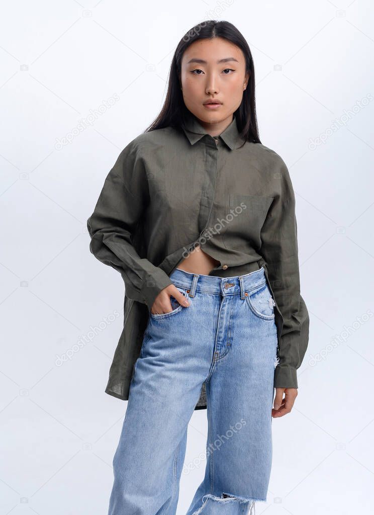 Beautiful Asian girl in a green linen shirt and casual jeans posing against a white wall in a photo studio