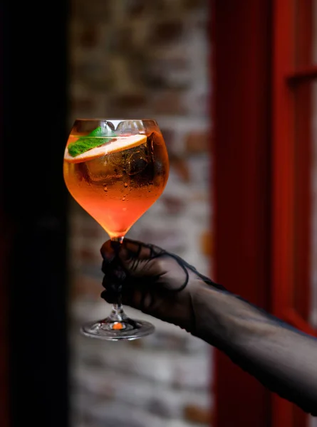 A female hand in black gloves holds a glass of Aperol cocktail on the background of the bar door