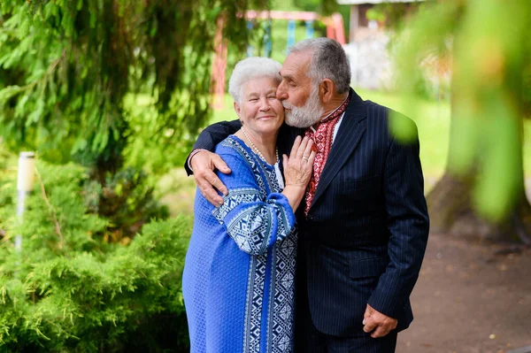 An old gray-haired woman hugs an old gray-haired man and sweetly kisses on the background of a park in Ukrainian traditional clothes