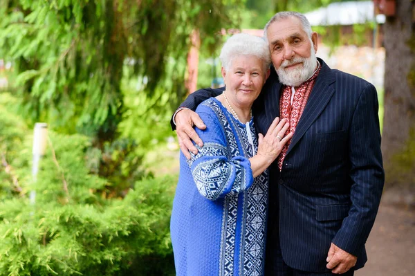 An old gray-haired woman hugs an old gray-haired man against the background of a park in Ukrainian traditional clothes