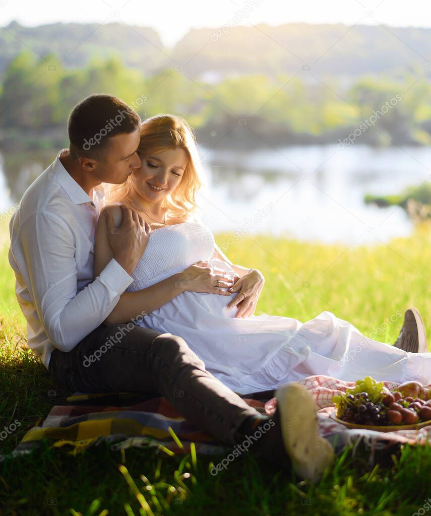 Beautiful young pregnant woman with her husband in the forest at sunset on a picnic sitting on the grass