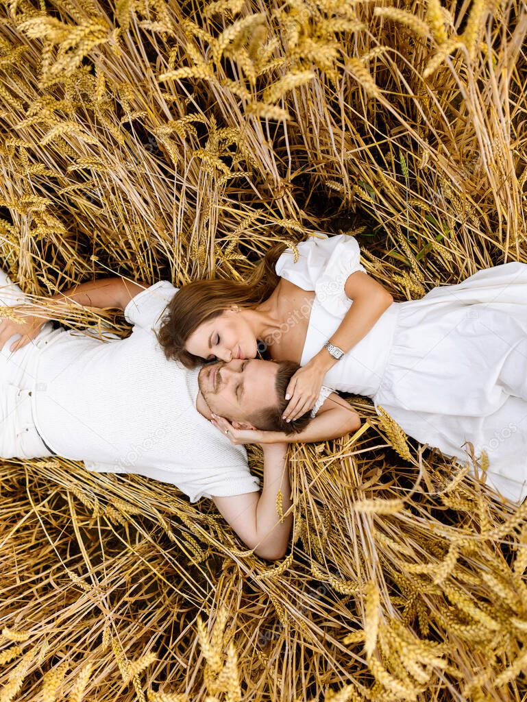A beautiful young Caucasian couple in white clothes are lying and hugging in a wheat field at sunset