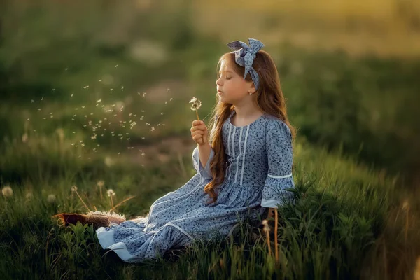 Mavi elbiseli bir kız çimlerin üzerinde oturur ve bir karahindiba tutar. Yaz.