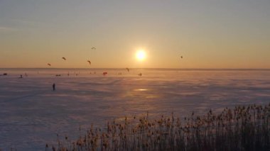 Donmuş bir kış nehrindeki bir grup sporcu, gün batımının arka planında kar uçurtmasıyla, renkli paraşütlerle ve insanların spor aktiviteleriyle hevesle meşguller. Pazar günleri eğleniyorlar. 4k 
