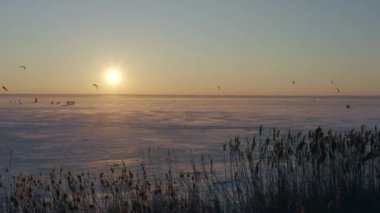 İnsanlar donmuş gölün buzlu bölgesinde kış sporlarıyla eğleniyorlar, akşamları gün batımında. Buzlu suda kar uçurtma, renkli paraşütlerin kontrolü. Buz yürüyüşü. Gökyüzünün altın gölgeleri. 4k görüntü