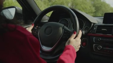 4K görüntülerde kırmızı kazaklı bir kız araba kullanıyor. Kadın ellerini direksiyondan ayırmıyor ve pencerenin dışında yeşil ağaçlar var. Dişinin ellerinde dövmeler var. Siyah arabanın içi..