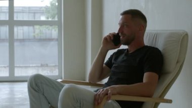 4K görüntü orta yaşlı bir adam telefonda mutlu bir şekilde konuşuyor rahat koltukta otururken gülümsüyor. Adam vakaları akıllı telefonla çözüyor. Hoş, büyüleyici bir sohbet. Önemli şeyler hakkında sakin bir diyalog.