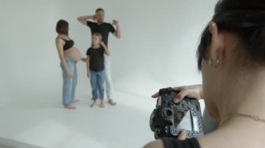 4K görüntülü fotoğrafçı kız fotoğraf çekimi sırasında kamerada fotoğraf çekiyor. Hamile bir anne, baba ve kızı beyaz bir arka planda poz vermeye hazırlanıyor.
