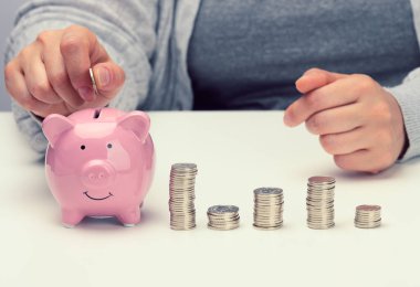 Kadın eli, beyaz bir masanın üzerindeki pembe kumbaraya bozuk para atıyor. Para biriktirme, tasarruf, sübvansiyon alma kavramı