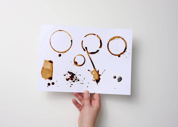 female hand holds a white sheet of paper with brown prints from the bottom of a cup with coffee, blots and splashes