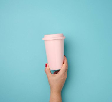 Mavi arka planda plastik kapaklı pembe bir seramik bardak tutan bir kadın el, mola zamanı ve kahve içmek