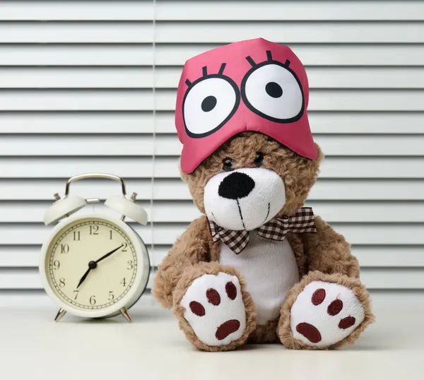 棕色的泰迪熊带着绷带坐在白桌子上睡觉 在闹钟附近 时间是早上7点 早上起来很困难 — 图库照片