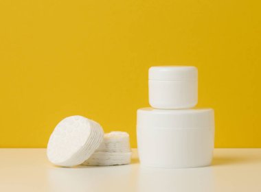 Beyaz plastik bir kavanozda kapağı ve beyaz pamuğu olan kozmetik ürünler. Marka ürünleri için boş, sarı arka planda nemlendirici.
