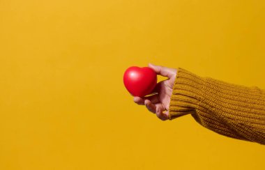 Kadın eli sarı arka planda kırmızı bir kalp tutuyor..