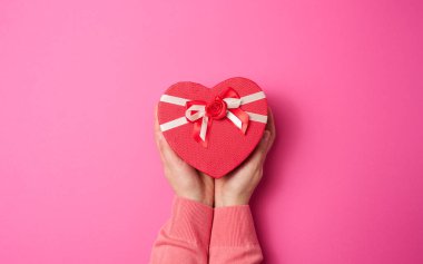 İki kadın eli pembe arka planda kırmızı kalp şeklinde bir kutu tutuyor. Şenlikli arkaplan