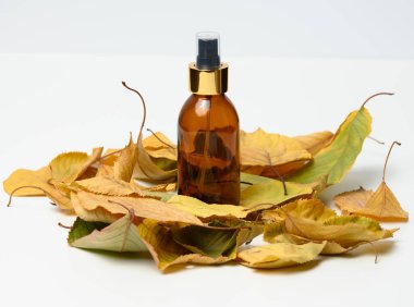 Sarı yaprakların ortasında beyaz arka planda kozmetik spreyli cam kahverengi şişe.