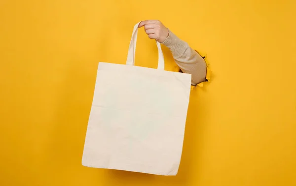 女性的手将一个空的白色纺织品袋放在黄色的背景上 身体的一部分从纸背的撕破的孔里伸出来 可重复使用和可循环利用的包装 无塑料 — 图库照片