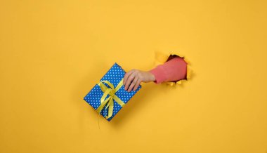 Kadın eli sarı arka planda bir hediye kutusunu tutuyor, vücudun bir parçası yırtık bir delikten kağıt bir arka planda çıkıyor. Tebrikler, tatil sürprizi.