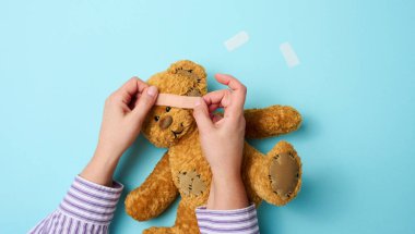 Kadın eli kahverengi bir oyuncak ayıyı tutuyor ve mavi bir arka planda tıbbi yapıştırıcı yapıştırıyor.