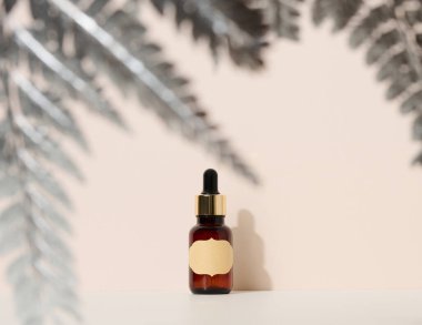 Kahverengi arka planda pipetli kahverengi cam şişe duruyor. Kozmetik, serum ve yağ için konteynır, gümüş eğreltiotu yaprağı