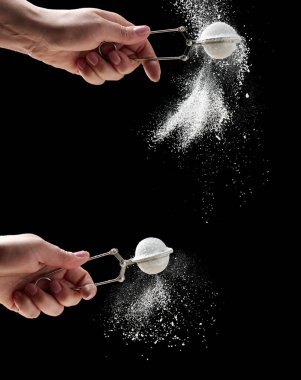 Kadın eli pudra şekerli metal bir süzgeç tutuyor. Parçacıklar siyah bir arkaplanda farklı yönlere uçarlar.