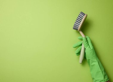 Yeşil lastik bir eldiven içinde yeşil bir arka planda plastik bir temizlik fırçası tutuyor.