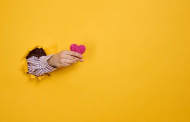 Kadın eli sarı arka planda kırmızı bir kalp tutuyor. Vücudun bir parçası yırtılan kağıttan fırlıyor.