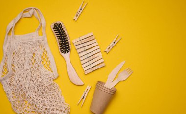 Beyaz pamuk torbası, kağıt bardaklar ve sarı arka planda tahta çatallar ve kaşıklar. Geri dönüştürülebilir atık, üst görünüm