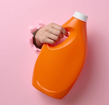 Pembe arka planda kadın elinde sıvı deterjan bulunan turuncu plastik şişe. Vücudun bir parçası arka plandaki yırtık delikten dışarı fırlıyor.