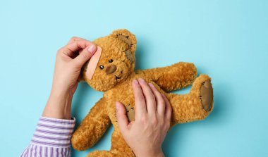 Kadın eli kahverengi bir oyuncak ayıyı tutuyor ve mavi bir arka planda tıbbi yapıştırıcı yapıştırıyor.