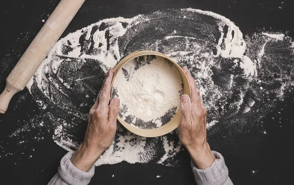 Kadın eli, siyah bir masanın üzerinde beyaz buğday unuyla yuvarlak bir ahşap elek tutar.
