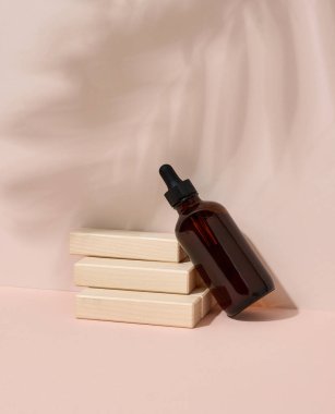 Kozmetik ürünleri, yağlar ve ahşap basamaklı bej arkaplanda serumu olan kahverengi cam bir şişe. Arka planda bir palmiye yaprağının gölgesi