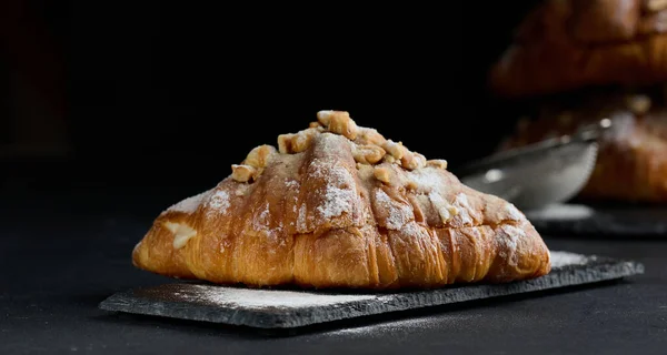 Baked croissant on a wooden board and sprinkled with powdered sugar, black table. Appetizing pastries for breakfast