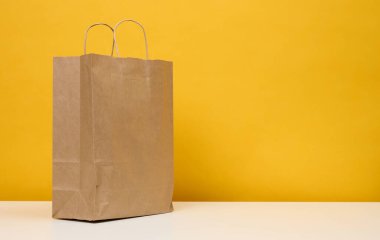 A brown paper bag stands on a white table, yellow background. Product packaging, copy space