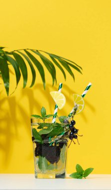 Transparent glass with lemonade, mint leaves, lemon slices and blackberries in the middle. Yellow background